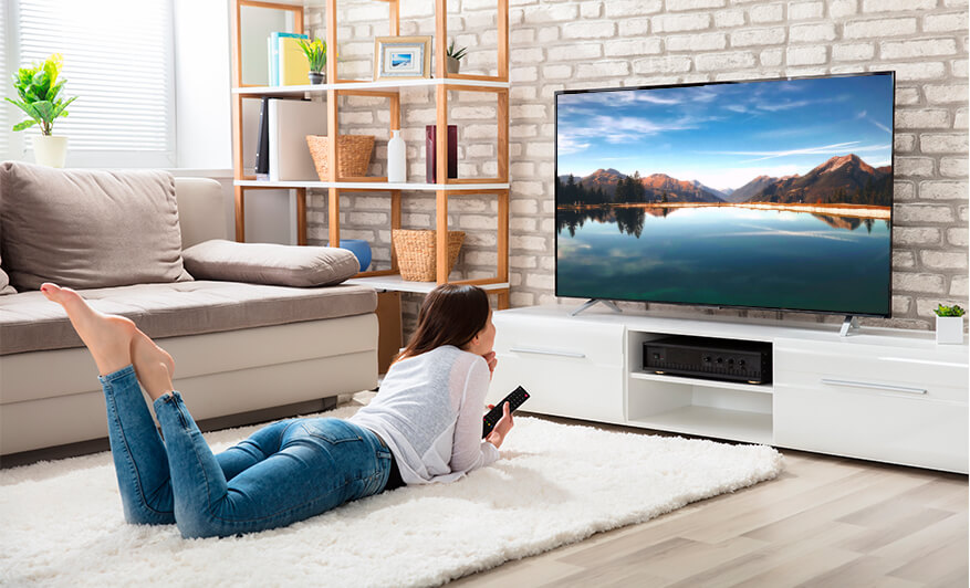Brunette female laying on fluffy white carpet in neutral  interior living room, watching TV 